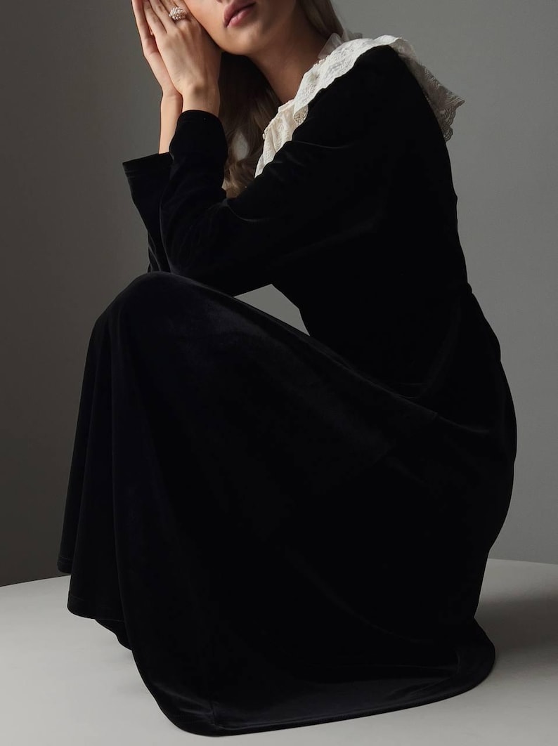 Pu&ograve; includere: Abito lungo nero in velluto con colletto in pizzo bianco. L'abito ha maniche lunghe e una gonna fluida. La modella &egrave; seduta, con le mani vicino al viso. L'abito &egrave; uno stile classico ed elegante.