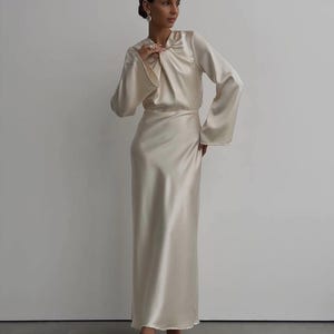 May include: A cream-colored satin dress with long sleeves and a floor-length skirt. The top features a draped neckline and the sleeves flare out from the elbow. The dress is worn with matching heels.