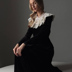 Pu&ograve; includere: Un lungo abito nero di velluto con un colletto alto in pizzo arricciato. L'abito ha maniche lunghe e una gonna fluida. La modella &egrave; in ginocchio, con i capelli biondi e un anello al dito. Lo sfondo &egrave; grigio.
