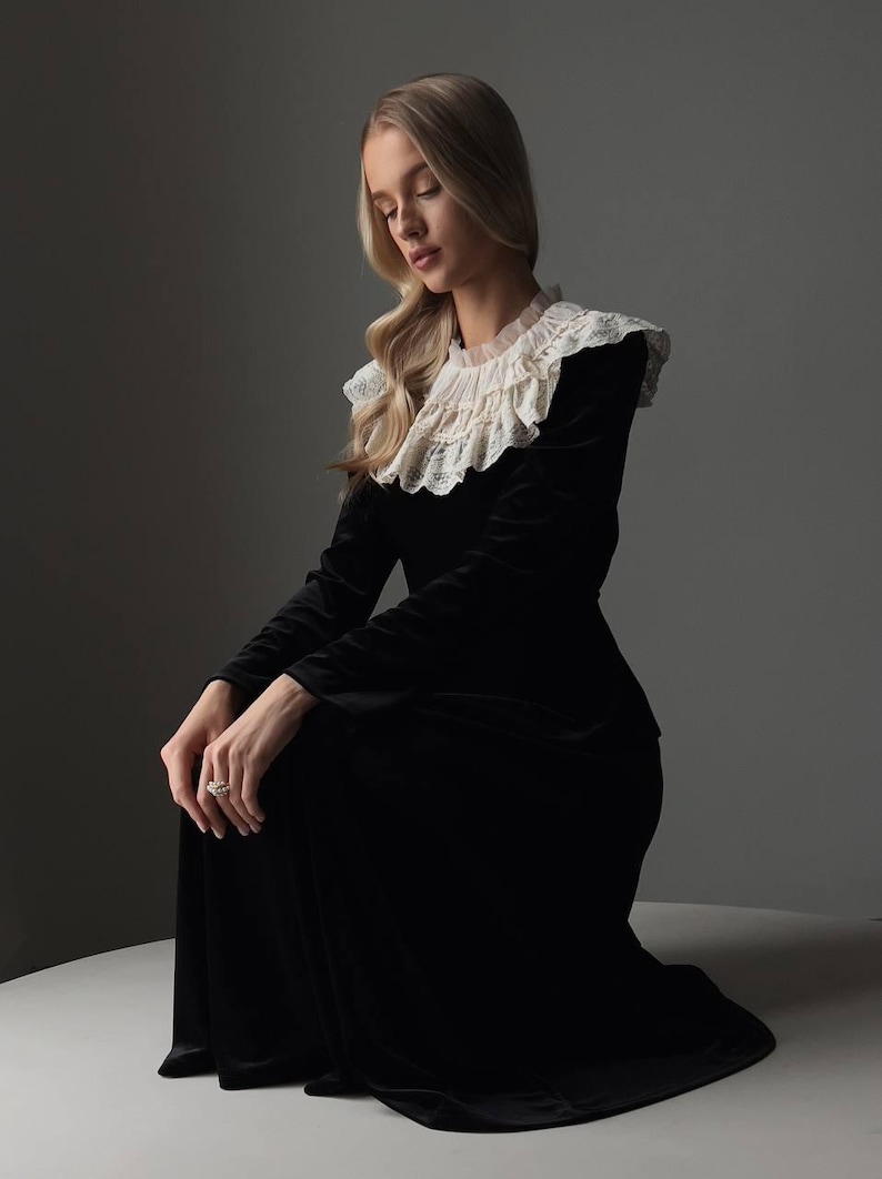 Pu&ograve; includere: Un lungo abito nero di velluto con un colletto alto in pizzo arricciato. L'abito ha maniche lunghe e una gonna fluida. La modella &egrave; in ginocchio, con i capelli biondi e un anello al dito. Lo sfondo &egrave; grigio.