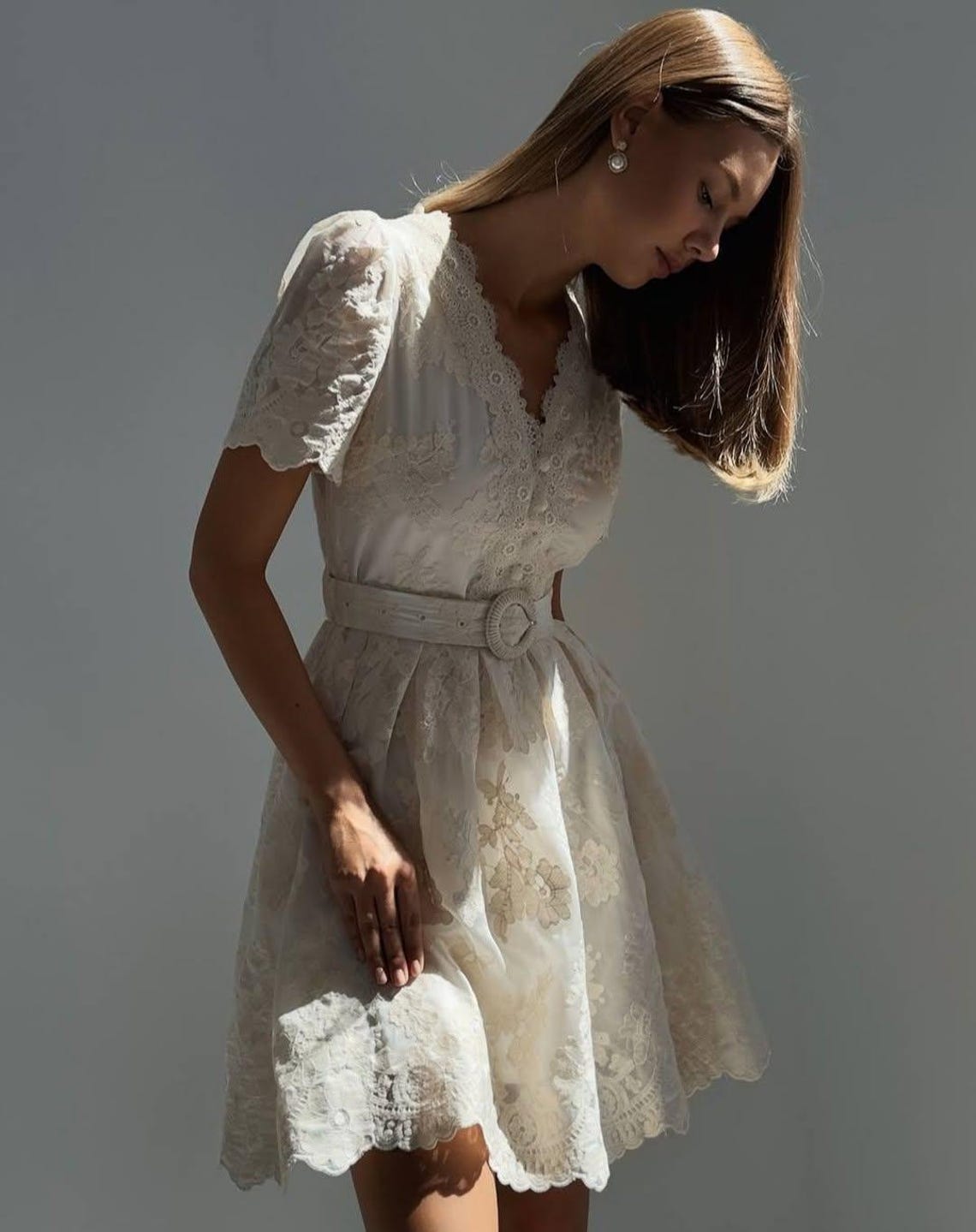 May include: A white, short-sleeved dress with a lace overlay and a floral pattern. The dress has a V-neckline, a belt with a buckle, and a scalloped hem.