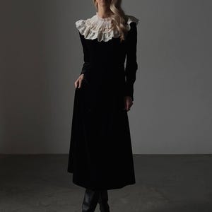 Pu&ograve; includere: Un lungo abito nero in velluto con colletto in pizzo bianco. L'abito ha maniche lunghe e una gonna svasata, abbinato a stivali neri al ginocchio. L'ambientazione &egrave; uno studio con illuminazione soffusa.