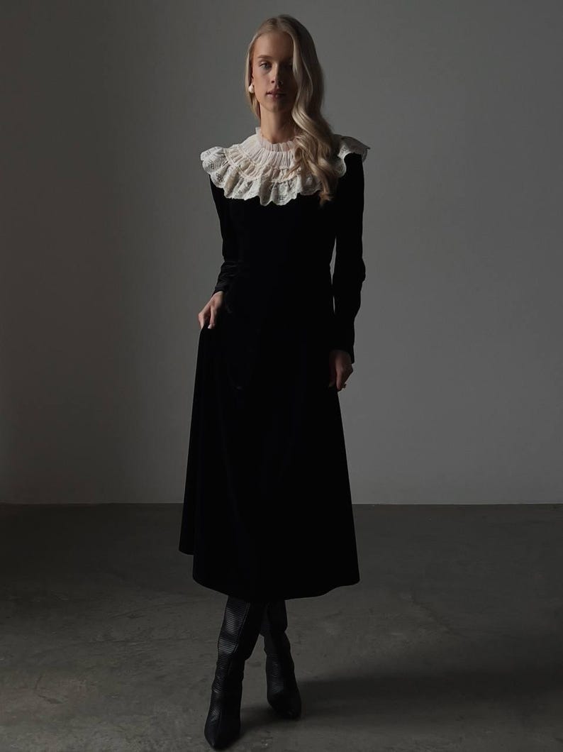 Pu&ograve; includere: Un lungo abito nero in velluto con colletto in pizzo bianco. L'abito ha maniche lunghe e una gonna svasata, abbinato a stivali neri al ginocchio. L'ambientazione &egrave; uno studio con illuminazione soffusa.