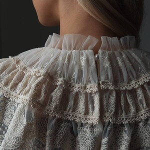 Pu&ograve; includere: Primo piano di un colletto in pizzo color crema con pi&ugrave; strati a balze. Il colletto presenta intricati motivi floreali e bordi delicati. Un bottone di perla chiude il colletto sul retro. Accessorio per il collo di ispirazione vintage.