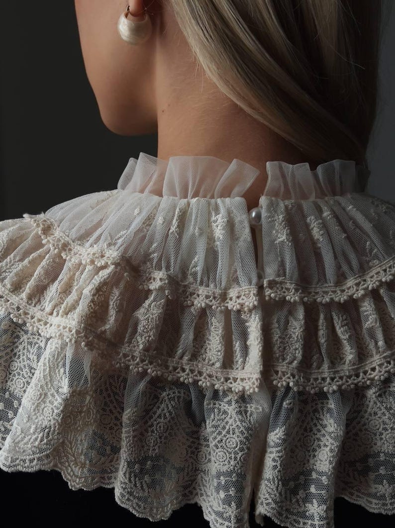 Pu&ograve; includere: Primo piano di un colletto in pizzo color crema con pi&ugrave; strati a balze. Il colletto presenta intricati motivi floreali e bordi delicati. Un bottone di perla chiude il colletto sul retro. Accessorio per il collo di ispirazione vintage.