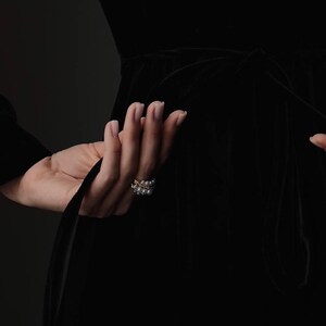 Pu&ograve; includere: Una mano con un anello d'oro con perle, che tiene un tessuto nero. L'anello ha un design a doppia fascia con piccole perle. Lo sfondo &egrave; di colore scuro e uniforme.
