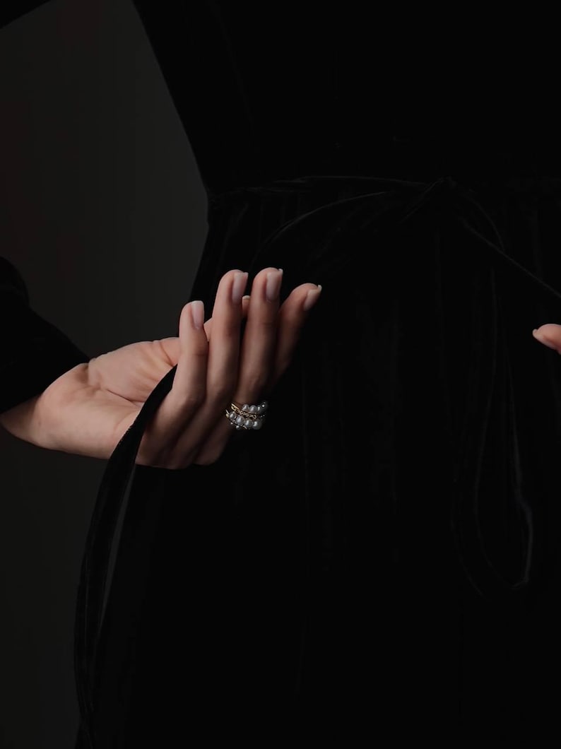 Pu&ograve; includere: Una mano con un anello d'oro con perle, che tiene un tessuto nero. L'anello ha un design a doppia fascia con piccole perle. Lo sfondo &egrave; di colore scuro e uniforme.
