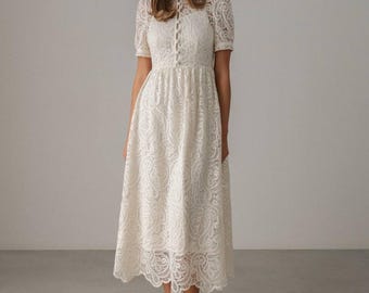 Può includere: Abito in pizzo color avorio con maniche corte e collo alto. L'abito presenta un'abbottonatura frontale e una gonna svasata, creando una silhouette elegante. L'intricato motivo in pizzo aggiunge una texture delicata al capo. L'abito è abbinato a scarpe color avorio abbinate.