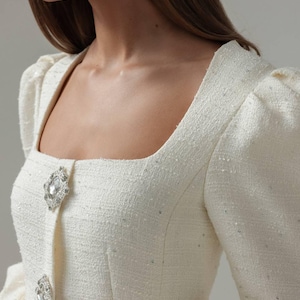 Könnte beinhalten: Cremefarbene Tweedjacke mit quadratischem Ausschnitt und langen Ärmeln. Die Jacke hat zwei große, verzierte Strassknöpfe und dezente Paillettenverzierungen. Die Ärmel haben ein Puffdesign an den Schultern.