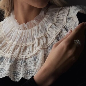 Pu&ograve; includere: Colletto in pizzo color crema con pi&ugrave; strati di balze e delicati motivi floreali. Il colletto &egrave; abbinato a un capo nero. &Egrave; visibile un anello di perle.