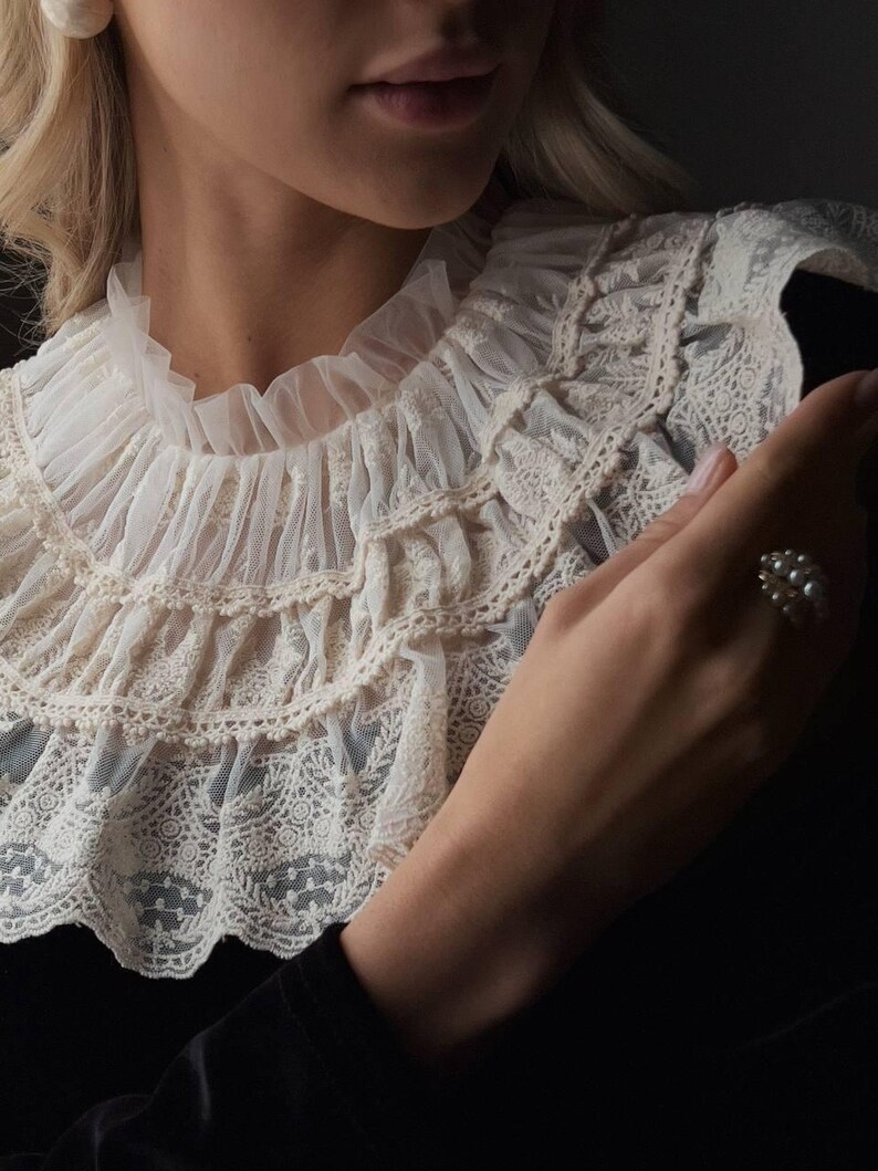 Pu&ograve; includere: Colletto in pizzo color crema con pi&ugrave; strati di balze e delicati motivi floreali. Il colletto &egrave; abbinato a un capo nero. &Egrave; visibile un anello di perle.