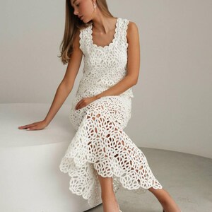 May include: A white crochet dress with a sleeveless top and a scalloped edge. The dress has an open, geometric design and is worn with white strappy sandals. The outfit is suitable for a summer event or special occasion.