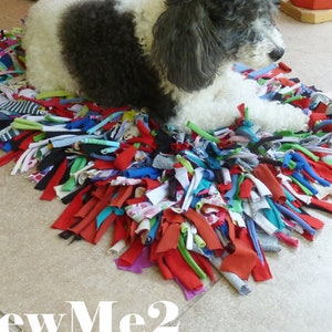 May include: A small, black and white dog lays on a colorful, handmade rag rug. The rug is made from strips of fabric tied together. The dog is looking to the right of the image.