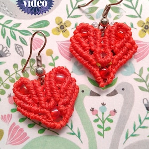 以下が含まれることがあります： 手作りの赤いマクラメのハート型イヤリングのペア。ビーズのアクセント付き。イヤリングは銅色のイヤーフックから吊り下げられています。背景には花柄と白鳥のパターンがあります。「USewMe2」の文字が下にあります。