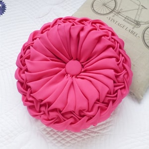 May include: A bright pink round pillow with a button in the center and a ruffled design. The pillow is on a white surface.