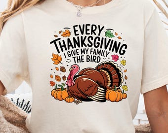 Może przedstawiać: Kremowy t-shirt z krótkim rękawem z grafiką brązowego indyka otoczonego pomarańczowymi dyniami, żołędziami i kolorowymi jesiennymi liśćmi. Napis brzmi "EVERY THANKSGIVING I GIVE MY FAMILY THE BIRD" czarnymi, pogrubionymi literami.