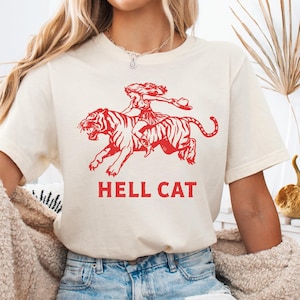 Könnte beinhalten: Cremefarbenes T-Shirt mit roter Grafik einer Frau, die auf einem Tiger reitet, und dem Text "HELL CAT" darunter. Das Design ist eine kühne, von Vintage inspirierte Illustration. Das Shirt ist aus weichem Material.