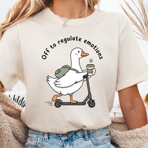 Puede incluir: Camiseta color crema con un dibujo de un ganso de dibujos animados montado en un patinete, sosteniendo una taza de café. El ganso lleva una mochila y el texto "Off to regulate emotions" está impreso encima. La camiseta se combina con pantalones cortos vaqueros.