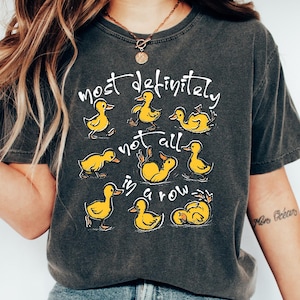 Puede incluir: Camiseta gris oscuro con la frase "most definitely not all in a row" e ilustraciones de patitos amarillos. La camiseta tiene un corte holgado y mangas cortas.