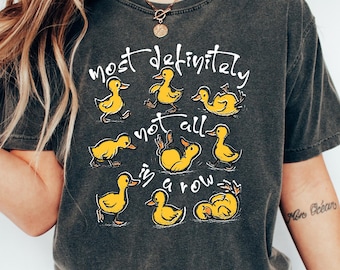 May include: A charcoal grey short-sleeve t-shirt featuring a graphic of nine yellow ducklings in various poses. The white text printed on the front reads, "most definitely not all in a row," arranged around the illustrated ducklings.