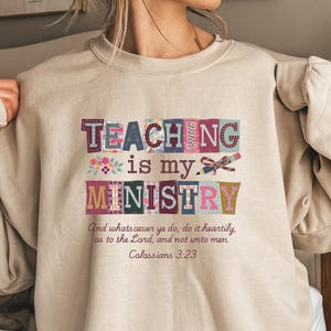 May include: Beige sweatshirt with the text "TEACHING is my MINISTRY" in colorful block letters. The sweatshirt also features the Bible verse Colossians 3:23. A comfortable and stylish top for teachers.