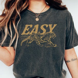 May include: Dark gray t-shirt with a distressed gold tiger graphic and the word "EASY" above it. The tiger is walking to the right. The shirt is a short-sleeved crew neck.