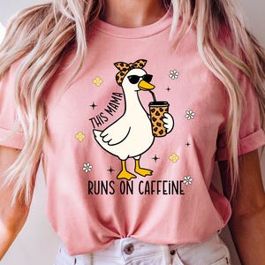 以下が含まれることがあります： サングラスとヒョウ柄のヘアバンドをつけたアヒルの漫画がコーヒーカップを持っているピンクのTシャツ。「THIS MAMA RUNS ON CAFFEINE」の文字と花のアクセントがプリントされています。