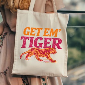 Peut inclure: Sac fourre-tout crème avec le texte "GET EM' TIGER" en orange et rose. En dessous, un dessin de tigre à rayures orange et rouge. Le sac est porté par une personne.