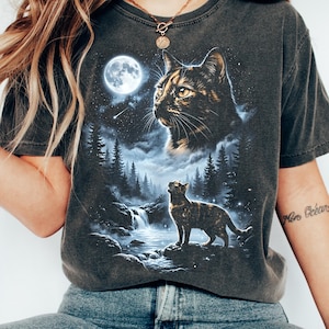 May include: A dark grey short-sleeve t-shirt featuring a graphic print of a tortoiseshell cat looking up at a full moon above a forest and waterfall. A second, larger cat head is positioned in the starry night sky above the trees.
