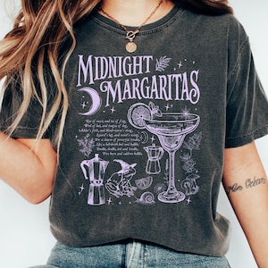 以下が含まれることがあります： ライトパープルのグラフィックプリントが施されたチャコールグレーの半袖Tシャツ。「MIDNIGHT MARGARITAS」の文字、マルガリータグラス、魔女の帽子をかぶったカエル、コーヒーポット、カタツムリ、植物のモチーフがデザインされています。