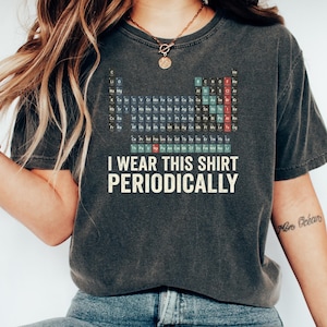 Op de afbeelding: Een antracietgrijs t-shirt met korte mouwen en een afbeelding van het periodiek systeem der elementen in gedempte kleuren. Onder de tabel staat de tekst I WEAR THIS SHIRT PERIODICALLY in een vet, gebroken wit, schreefloos lettertype.