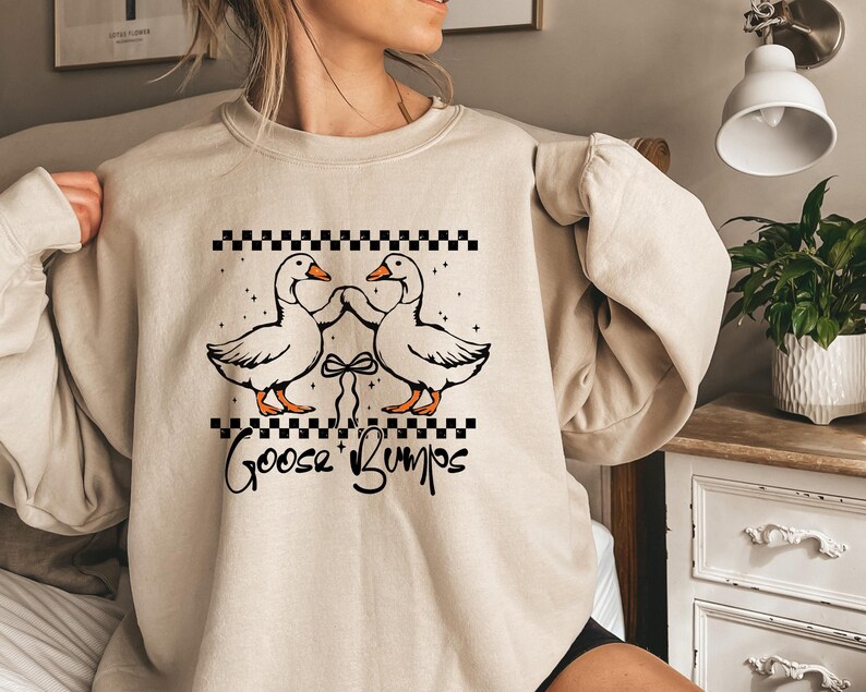Puede incluir: Sudadera beige con un gr&aacute;fico de dos gansos tomados de la mano, sobre un patr&oacute;n de cuadros blancos y negros. El texto "Goose Bumps" est&aacute; escrito debajo en una fuente de escritura. La sudadera tiene mangas largas.