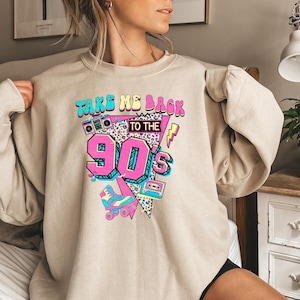May include: Beige sweatshirt with a colorful graphic design. The design features the text "TAKE ME BACK TO THE 90'S" in teal and pink, along with a boombox, roller skate, and cassette tape. The number "90's" is in a large, vibrant pink font.