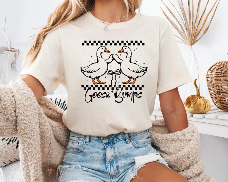 Puede incluir: Camiseta color crema con un gr&aacute;fico de dos gansos blancos tomados de la mano, con patas y picos naranjas, y un patr&oacute;n de cuadros blancos y negros arriba. El texto "Goose Bumps" est&aacute; escrito debajo de los gansos.