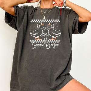 Puede incluir: Camiseta gris oscuro con un gr&aacute;fico blanco de dos gansos enfrentados, con un lazo entre ellos y las palabras "Goose Bumps" debajo. El dise&ntilde;o incluye un patr&oacute;n de cuadros encima y debajo de los gansos.