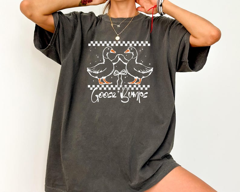 Puede incluir: Camiseta gris oscuro con un gr&aacute;fico blanco de dos gansos enfrentados, con un lazo entre ellos y las palabras "Goose Bumps" debajo. El dise&ntilde;o incluye un patr&oacute;n de cuadros encima y debajo de los gansos.