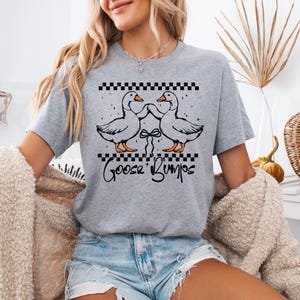 Puede incluir: Camiseta gris claro con un gr&aacute;fico de dos gansos enfrentados, con un lazo entre ellos. La camiseta tiene las palabras "Goose Bumps" en una fuente de escritura debajo de los gansos. La parte superior del dise&ntilde;o tiene un patr&oacute;n de cuadros blancos y negros.
