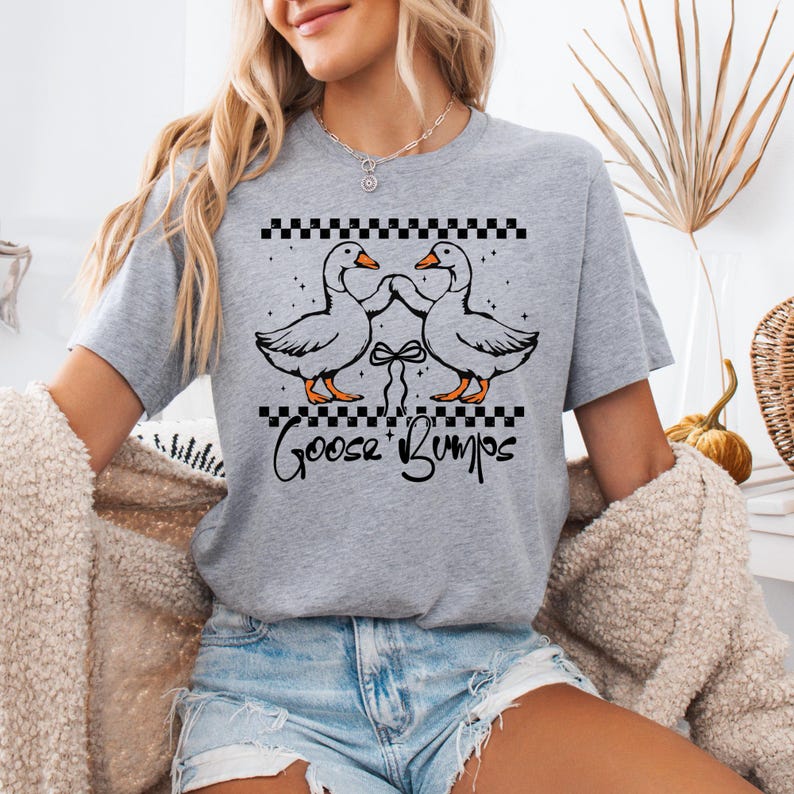 Puede incluir: Camiseta gris claro con un gr&aacute;fico de dos gansos enfrentados, con un lazo entre ellos. La camiseta tiene las palabras "Goose Bumps" en una fuente de escritura debajo de los gansos. La parte superior del dise&ntilde;o tiene un patr&oacute;n de cuadros blancos y negros.