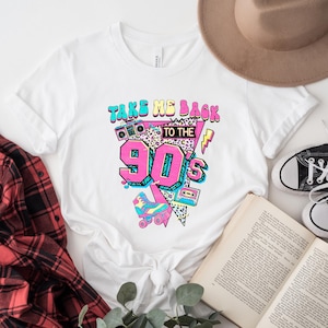 May include: White t-shirt with a colorful graphic design that says "Take Me Back to the 90's". The design includes a boombox, roller skates, and a cassette tape. A brown hat, a pair of black sneakers, and a book are also visible.