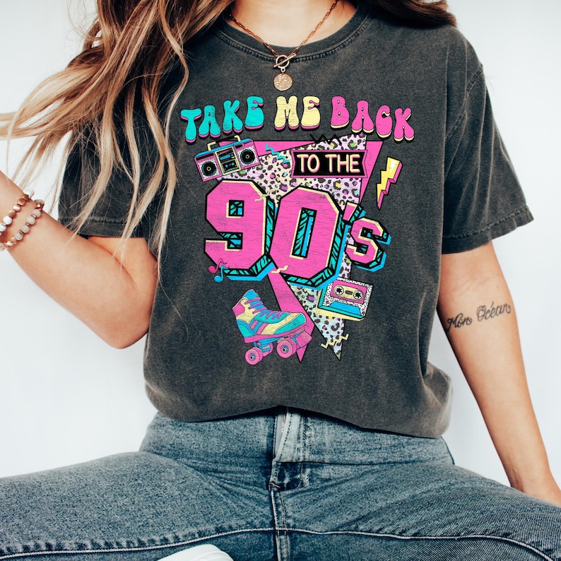 May include: Dark gray t-shirt with a colorful graphic that says "TAKE ME BACK TO THE 90'S". The graphic includes a boombox, roller skate, and cassette tape in bright colors. The shirt is worn with blue jeans.