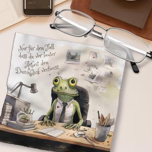 Könnte beinhalten: Ein quadratisches Mikrofasertuch mit einem grünen Frosch in weißem Hemd und Krawatte an einem Schreibtisch. Der Text lautet: Nur für den Fall, dass du vor lauter Arbeit den Durchblick verlierst. Das Tuch hat gezackte Ränder und liegt unter einer Brille mit silbernem Rahmen.