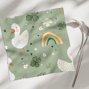 Könnte beinhalten: Ein quadratisches Stoffmuster mit hellgrünem Hintergrund, das mit weißen Gänsen, vierblättrigen Kleeblättern, kleinen weißen Blumen und einem Regenbogen gemustert ist. Die Gänse haben orangefarbene Schnäbel und Füße. Der Stoff hat einen Overlockrand.