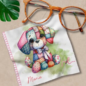 Könnte beinhalten: Ein weißes Mikrofaser-Reinigungstuch mit einer bunten Patchwork-Hundeillustration und dem Namen "Maria" darauf gedruckt. Das Tuch ist zum Reinigen von Brillen gedacht.