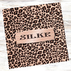 Könnte beinhalten: Ein quadratischer, beigefarbener Stoff mit Leopardenmuster. Ein Holzschild mit dem Wort "SILKE" in schwarzer Schrift liegt darauf. Der Stoff hat einen strukturierten, gefransten Rand.
