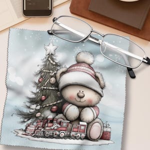 Könnte beinhalten: Ein hellblaues Mikrofasertuch mit einer weihnachtlichen Illustration. Das Design zeigt einen Teddybären mit rosa-weißem Hut, einen Weihnachtsbaum und eine Spielzeugeisenbahn. Eine Brille mit silbernem Rahmen liegt auf dem Tuch.