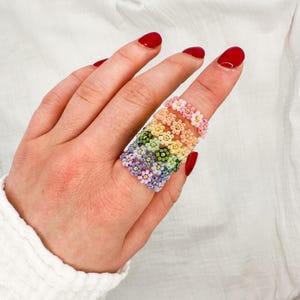 Op de afbeelding: Een hand met een verzameling bloemenringen van kralen in een regenboog van kleuren. De ringen zijn gemaakt van kleine kralen, waarbij elke ring een ander kleurenschema heeft, waaronder roze, oranje, geel, groen, blauw en paars.