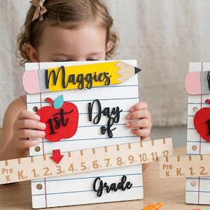 Może przedstawiać: Dwie białe drewniane tabliczki w kształcie kartki z notatnika, każda z żółtym ołówkiem, czerwonym jabłkiem z napisem 1st Day of Grade oraz grafiką linijki z liczbami od Pre-K do 12 i czerwoną strzałką.