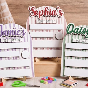Può includere: Tre insegne in legno ad arco con sfondi a righe in viola, rosa e verde. Ogni insegna presenta un nome in alto, seguito da campi di testo per First or Last Day Of, Today's Date, Age, Things I Love e When I Grow Up, I Want To Be.