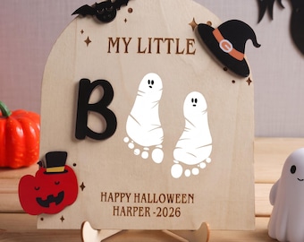 Könnte beinhalten: Ein gewölbtes Holzschild mit dem Text MY LITTLE, HAPPY HALLOWEEN HARPER -2026, einem schwarzen Buchstaben B, zwei weißen geisterförmigen Fußabdrücken, einer schwarzen Fledermaus, einem schwarzen Hexenhut und einem roten Kürbis auf einem Holzständer.