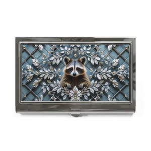 May include: A silver business card holder featuring a raccoon design. The raccoon is centered within a decorative silver leaf and vine pattern against a blue background. The holder has a silver frame and a clasp.